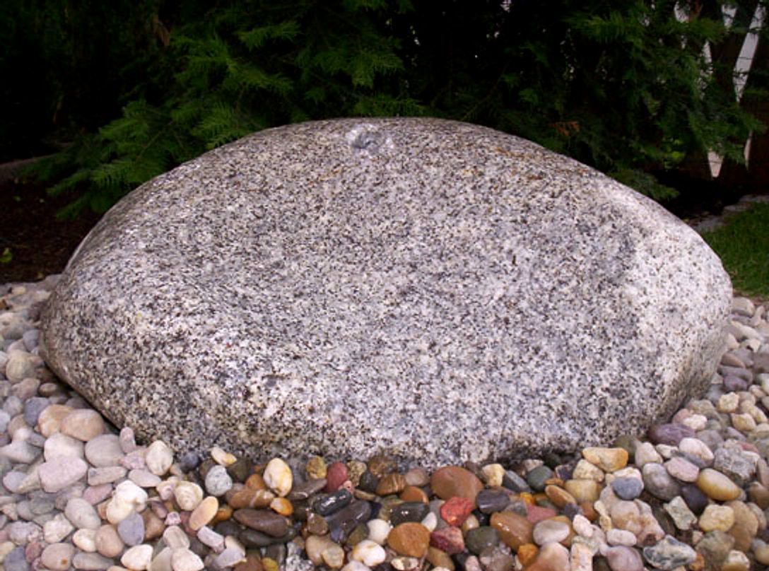 Grosser runder Granitfindling in einem Bett aus buntem Flusskies, dahinter dunkle Nadelgehoelze.
