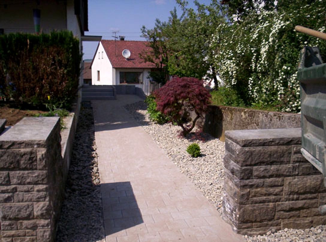 Schmaler Weg zwischen zwei Haeusern, gefasst von Mauerscheiben aus Beton, mit Kiesstreifen und einem roten Faecherahorn.