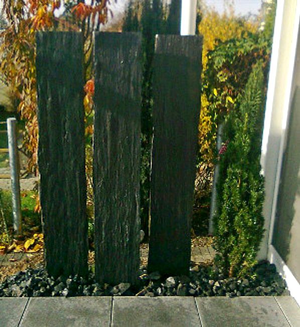 Vier hohe Stelen aus dunklem Schiefer stehen als Sichtschutz in einem Beet aus schwarzem Splitt, dahinter herbstlich gefaerbte Straeucher.