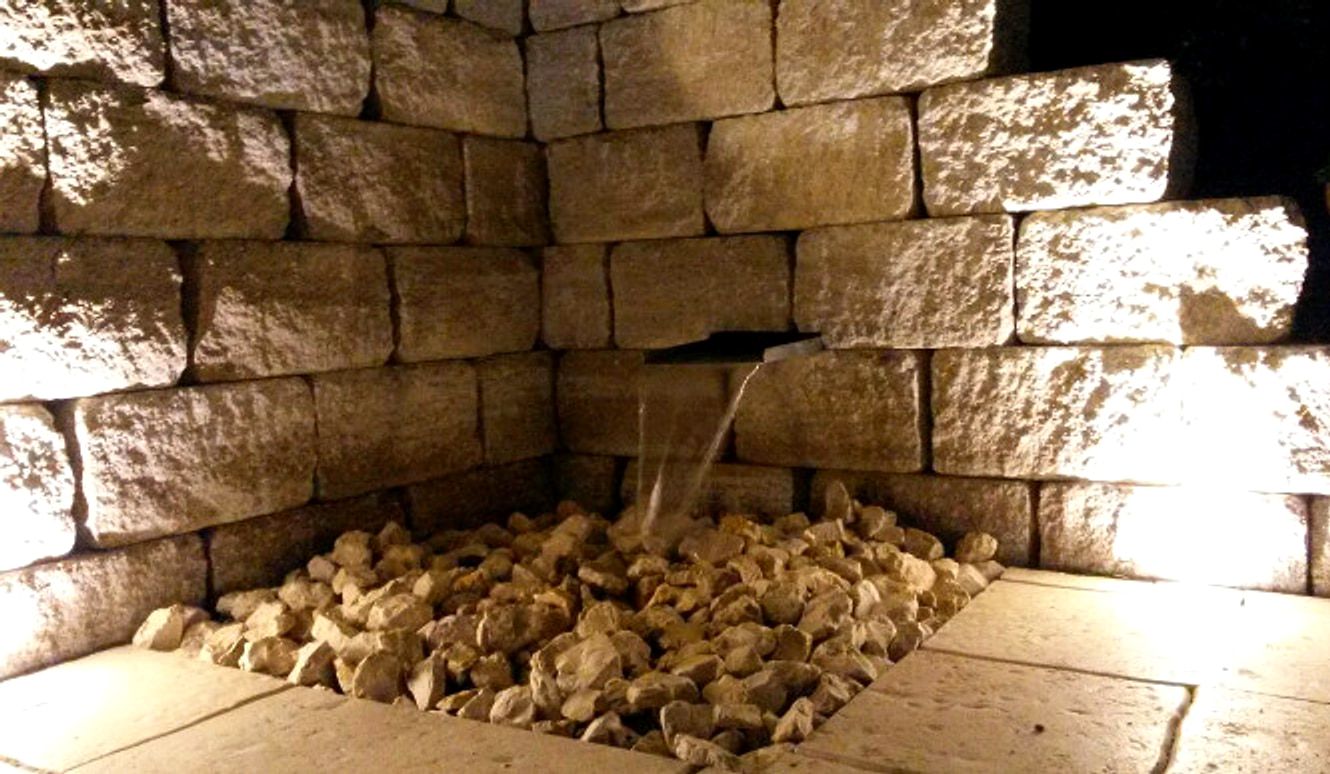 Beleuchtete Wasserwand bei Nacht: aus einer Muschelkalkmauer tritt ein schmaler Wasserstrahl und faellt auf ein Bett aus groben Bruchsteinen.
