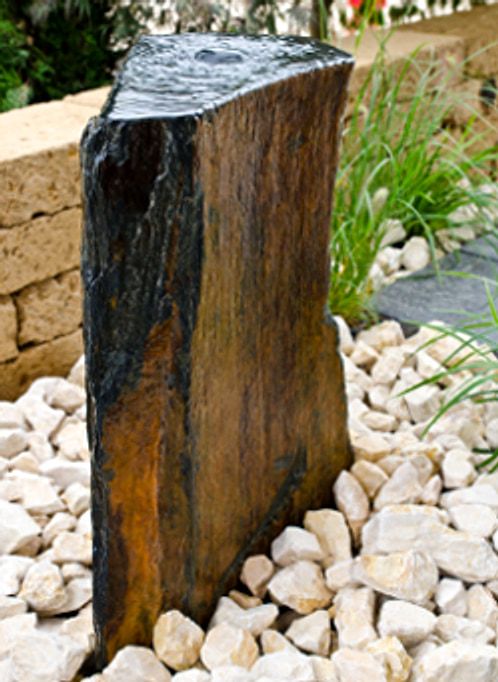 Derselbe Basalt-Quellstein von der Seite, das Wasser laeuft ueber die geriffelte Bruchkante in den hellen Kies.