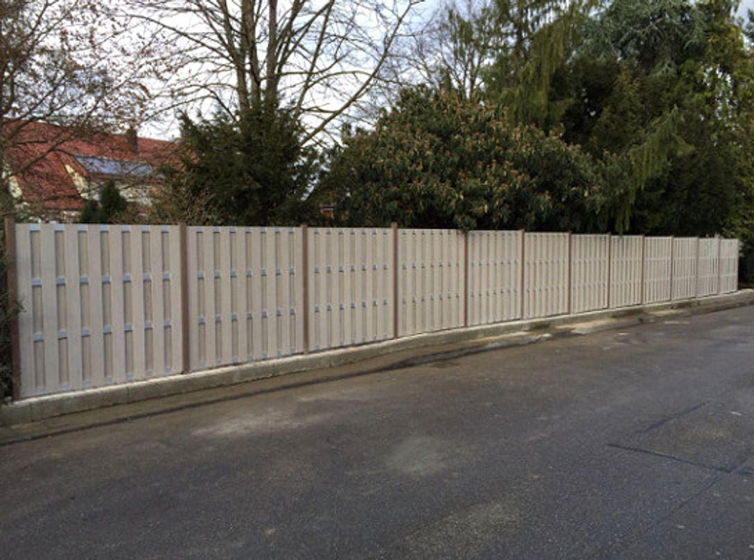Weisser Lattenzaun auf einer niedrigen Mauer entlang einer Strasse, dahinter hohe Nadelgehoelze.