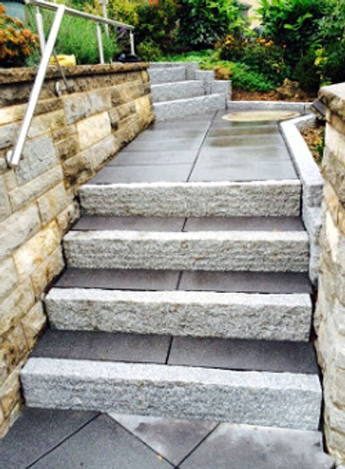 Treppe aus breiten Granitstufen zwischen zwei Muschelkalkmauern, mit Edelstahl-Handlauf und einer schmalen Wasserrinne im Zwischenpodest.