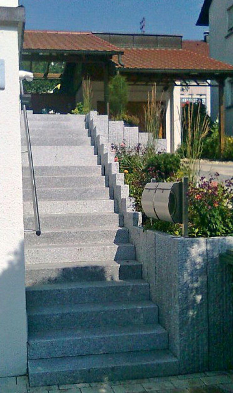 Steile Aussentreppe aus Granit-Blockstufen an einer Hauswand, rechts Granitpalisaden mit einem Staudenbeet und einem Briefkasten.